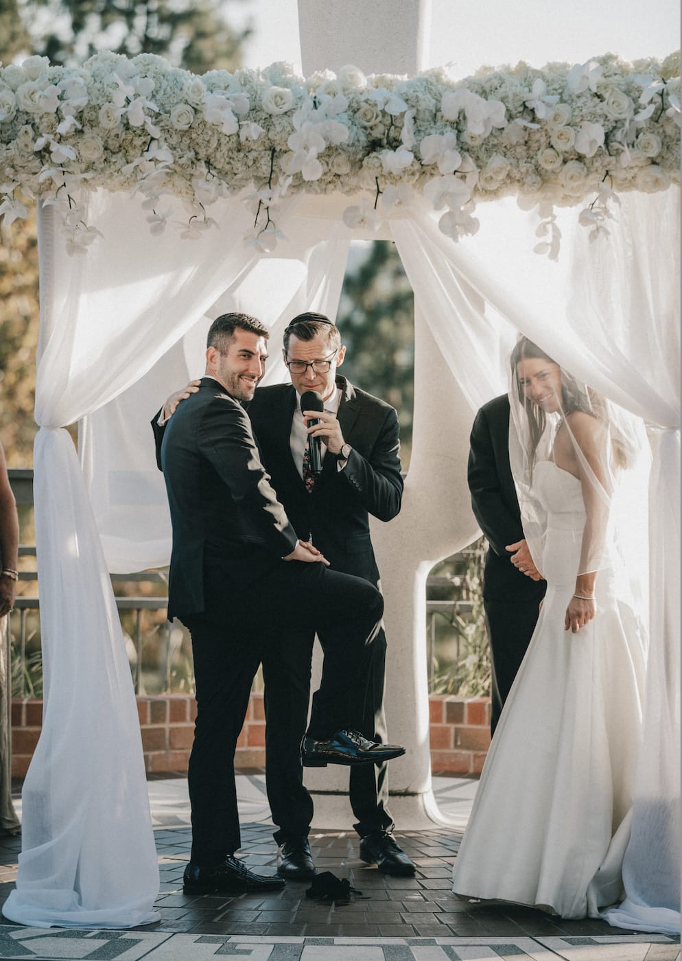 A couple under a white floral chuppah with Rabbi Mordechai Teller officiating